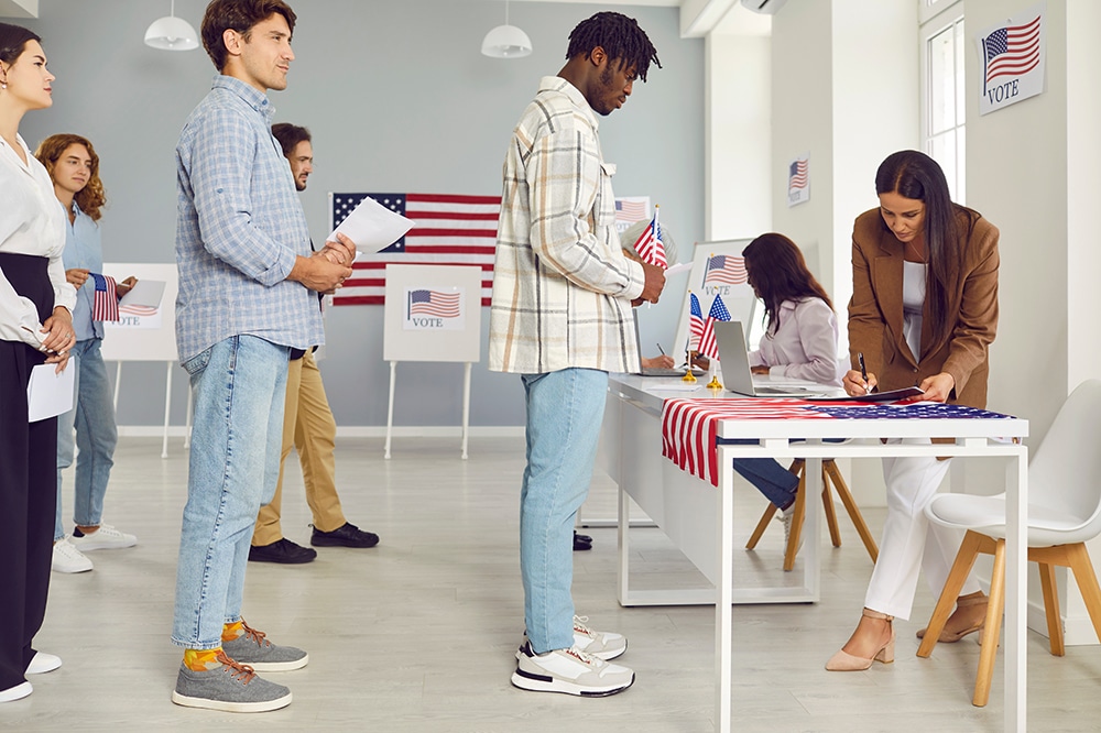 Voters lining up to vote at a polling location with a flag in the background.