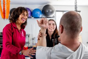 An NAU physical therapy student and a supervising physical therapy specialist smile at their patient, a man in a gray shirt, as the student adjusts his arm.