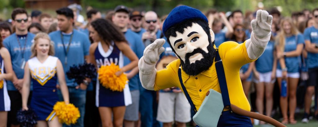 Louie with yellow shirt and overalls in front of cheer leading squad and other students with his arms up.