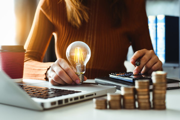 Woman holding a light bulb on top of a table that has a laptop and piles of coins