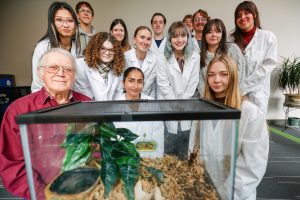 Thirteen research assistants in white lab coats and professor Larry Stevens pose around a glass enclosure containing a tarantula.