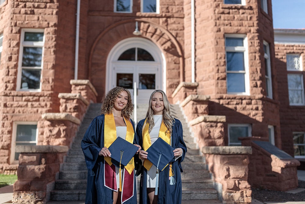 Justiss Green and Randi Garcia in graduation robes in front of Old Main