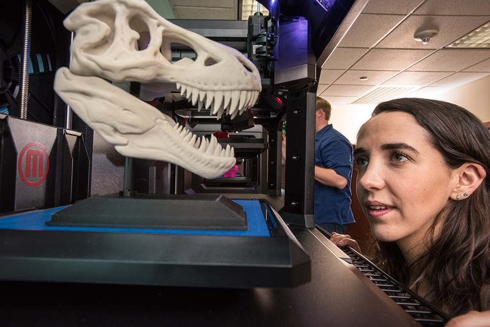 A person examining a large, dinosaur skull-shaped model on a 3D printer in a workshop setting.