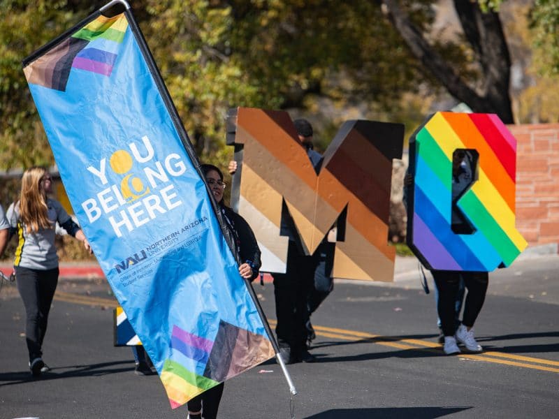 You Belong Here banner being carried in NAU parade next to letters painted with the skin tone rainbow and the pride rainbow